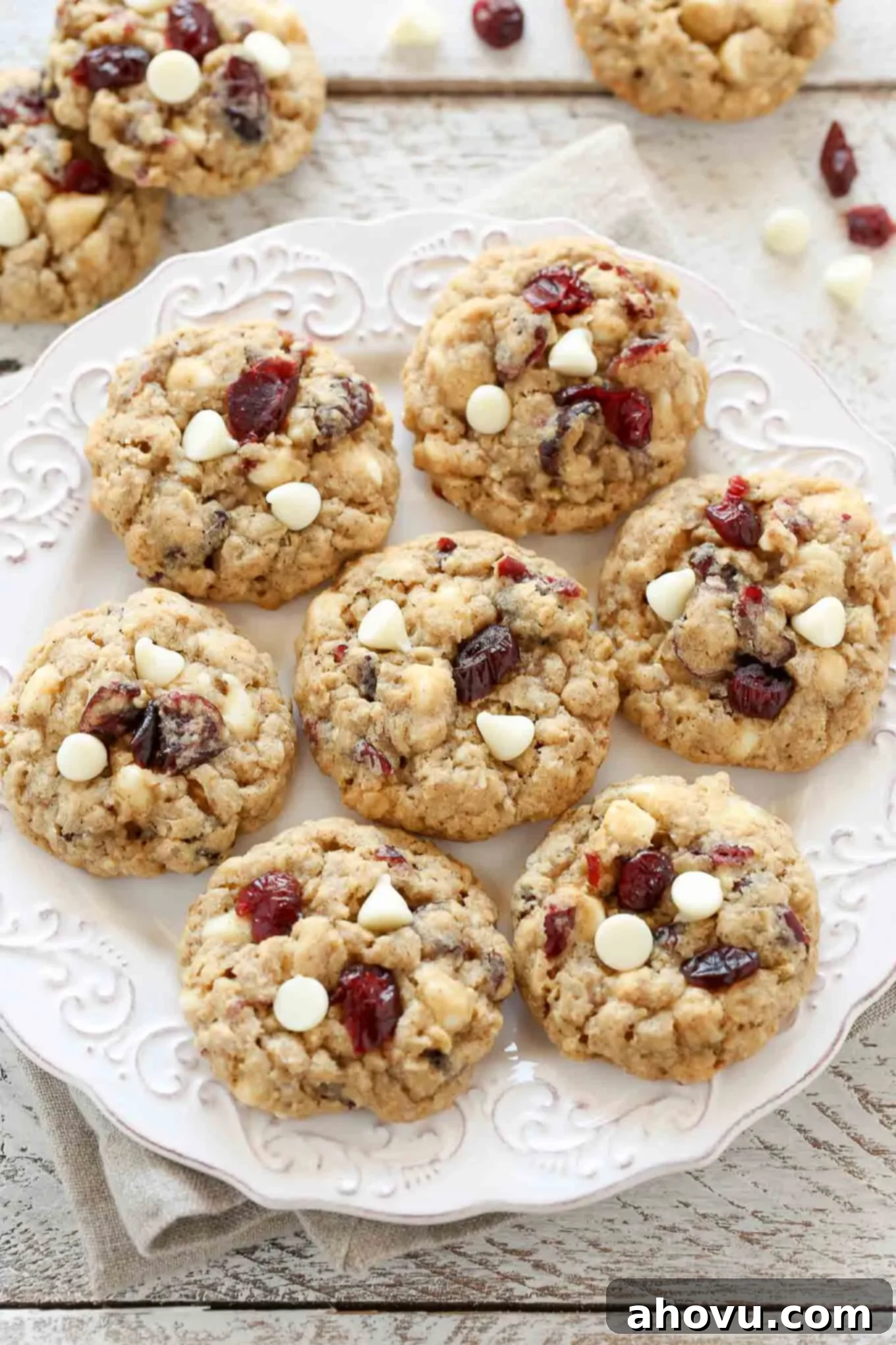Overhead view of seven soft and chewy white chocolate oatmeal cranberry cookies on a white plate, perfect for holidays. Additional cookies rest on a rustic counter next to a festive arrangement.
