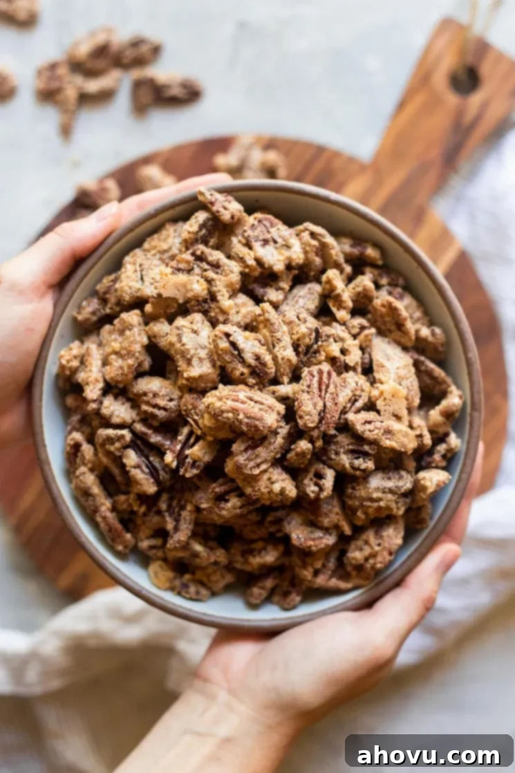 Sweet Crunchy Homemade Pecan Perfection 5 A bowl of freshly made candied pecans being gently held in the air over a round wooden tray and a soft striped linen, showcasing their golden, irresistible appeal.