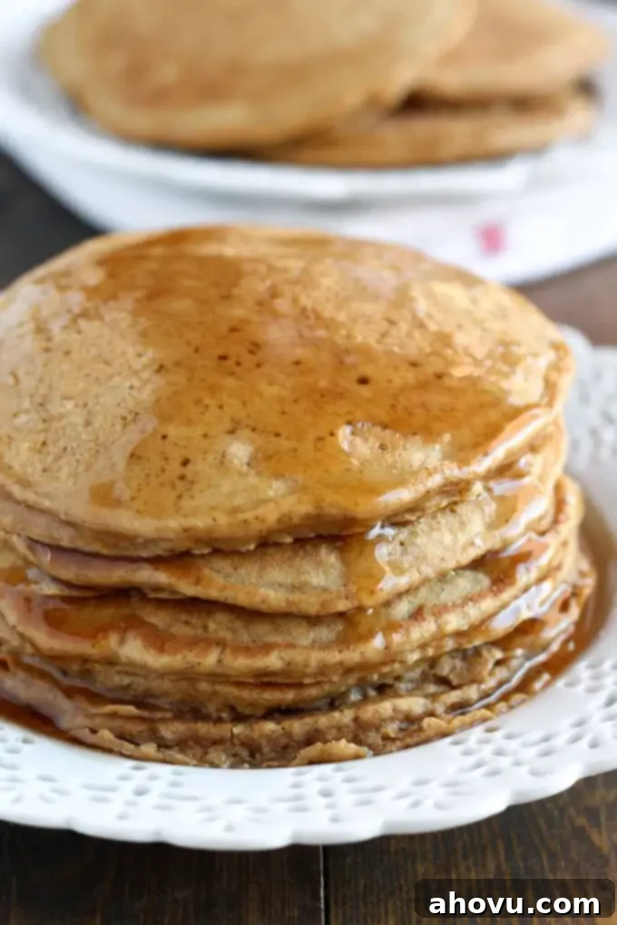 Cozy Gingerbread Flapjacks 3 An inviting close-up of a stack of gingerbread pancakes, artfully arranged on a pristine white plate. More freshly cooked pancakes are visible, softly blurred in the background, hinting at the abundance of this festive breakfast.