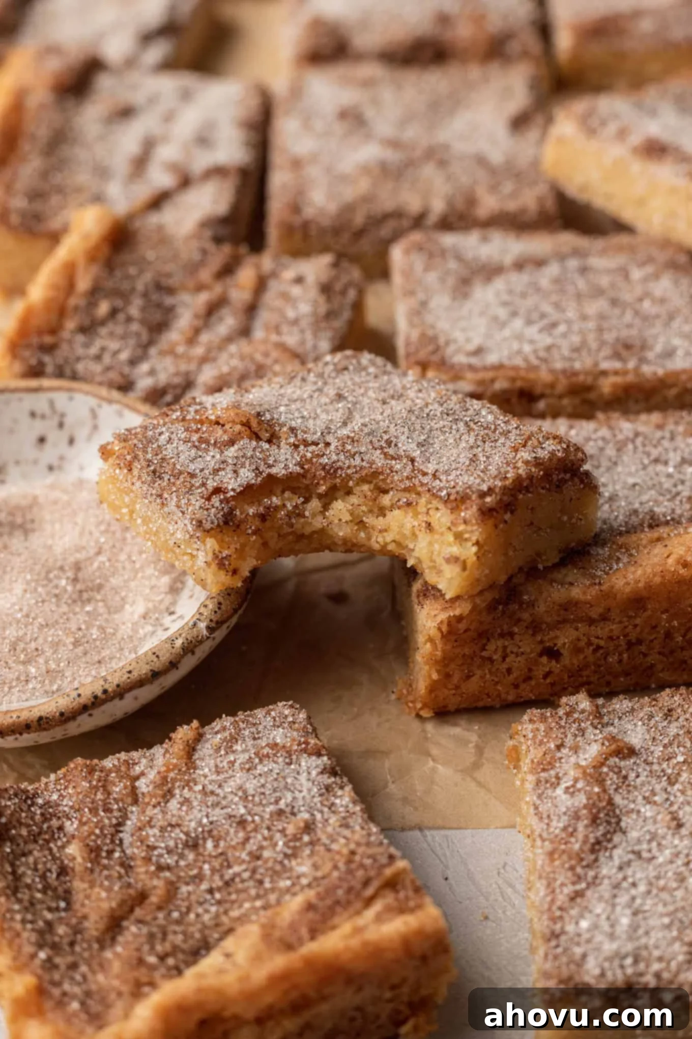 Delicious Snickerdoodle cookie bars on parchment paper. One bar has a bite missing and is resting atop the others, showcasing its soft interior and cinnamon-sugar topping.