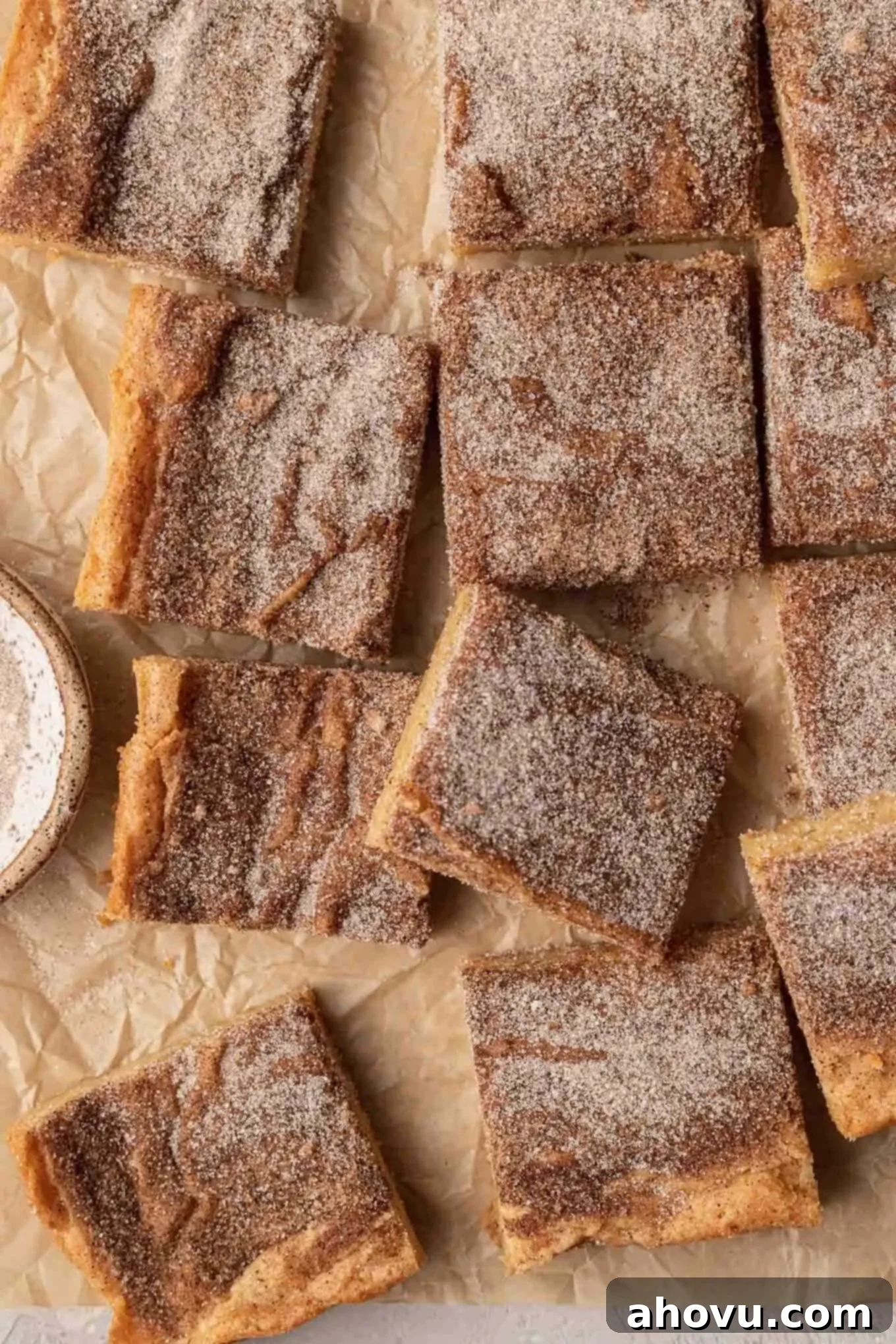An overhead view of freshly baked snickerdoodle blondies cooling on parchment paper, displaying their golden-brown tops and generous cinnamon-sugar coating.