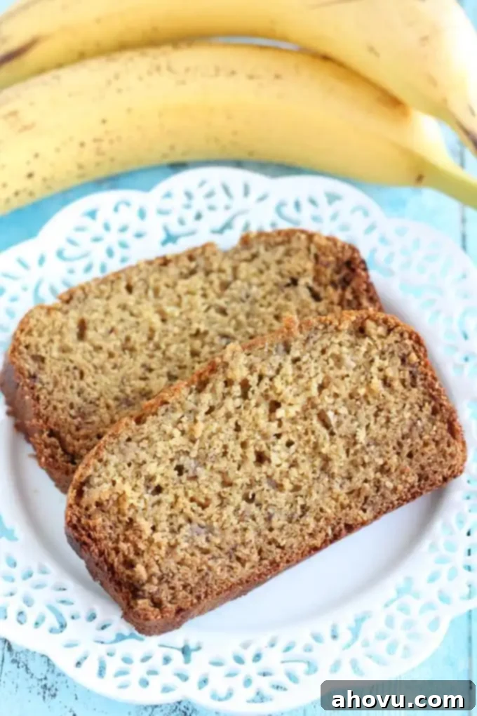 Two generous slices of moist whole wheat banana bread, garnished with a sprinkle of cinnamon, resting on a pristine white plate, with fresh, yellow bananas subtly blurred in the background.
