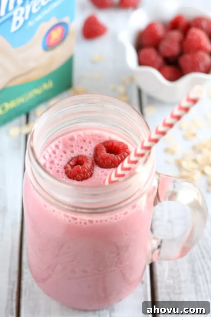 A raspberry smoothie in a glass mason jar with a striped straw. A carton of almond milk and a bowl of fresh raspberries rest in the background.
