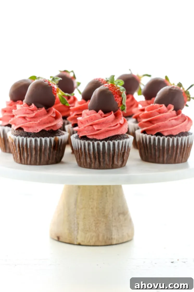 Chocolate cupcakes topped with strawberry buttercream frosting and chocolate covered strawberries on top of a white marble cake stand.