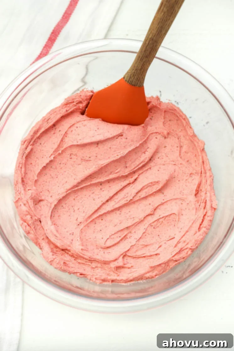 A glass bowl filled with strawberry buttercream frosting and a rubber spatula on the side.