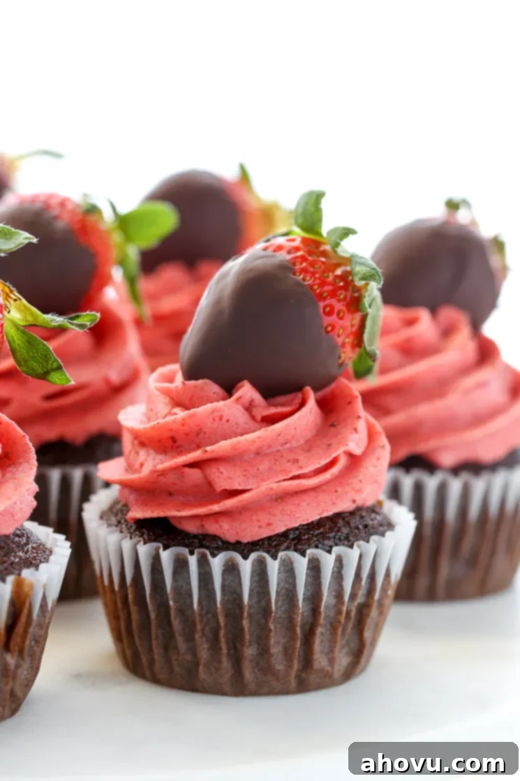 A closeup shot of chocolate cupcakes topped with strawberry buttercream frosting and chocolate covered strawberries on a white marble cake stand.