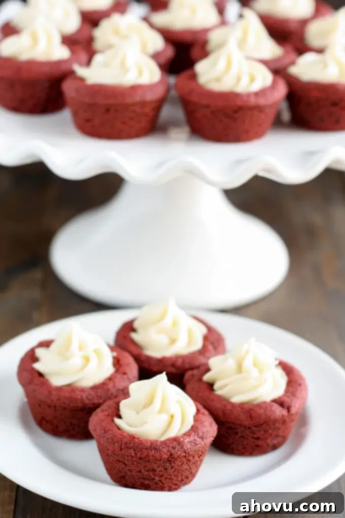 Red Velvet Cookie Bites 3 Deep dish red velvet cookie cups with cream cheese frosting on a white plate, with a cake stand of more cookie cups in the softly blurred background.