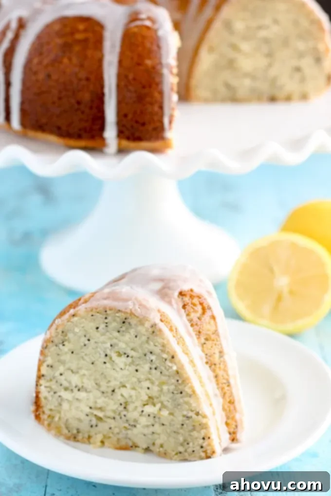 A beautiful slice of glazed lemon poppy seed bundt cake on a pristine white plate, showcasing its moist texture and delicate poppy seeds. In the background, the rest of the elegant bundt cake rests on a cake stand, glistening with lemon glaze.