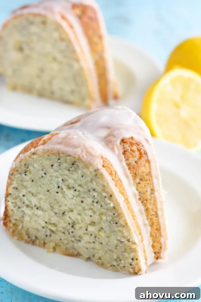 A perfectly glazed slice of lemon poppy seed cake rests on a white plate, ready to be enjoyed. In the soft-focus background, fresh lemon halves and another slice of the delicious cake are visible, hinting at the delightful citrus experience.