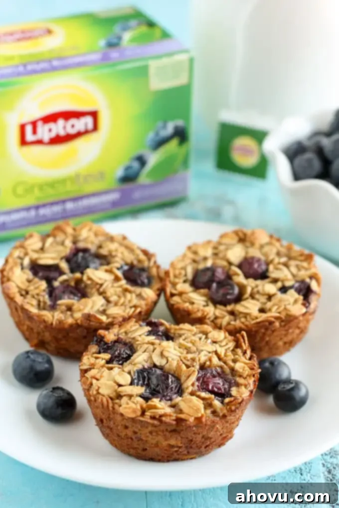 Berry Bliss Baked Oats 2 Three healthy blueberry baked oatmeal cups on a white plate with fresh berries and a sprig of mint. A box of Lipton tea and a bowl of fresh blueberries rest in the blurred background.