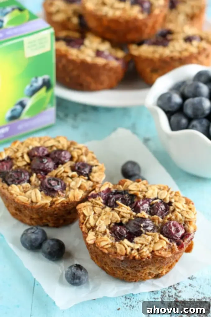 Berry Bliss Baked Oats 4 Two perfectly baked blueberry oatmeal cups, topped with fresh blueberries, resting on a piece of parchment paper. More oatmeal cups, a bowl of fresh berries, and a box of tea are in the soft background.