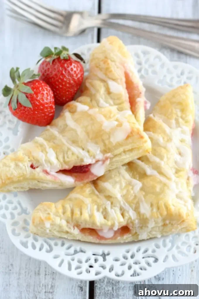 Two golden-brown strawberry turnovers on a decorative white plate, garnished with fresh strawberries. Two forks rest gently in the background, hinting at immediate enjoyment.