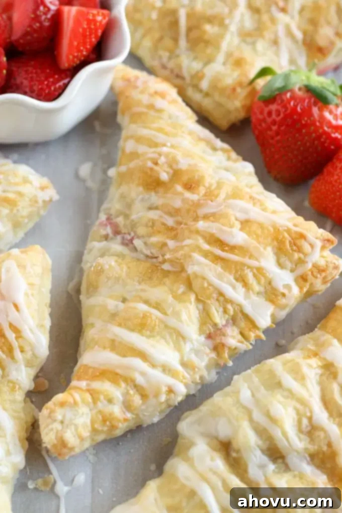 Several golden-brown strawberry turnovers resting on parchment paper, alongside a white bowl filled with fresh, sliced strawberries. This showcases the key ingredient.