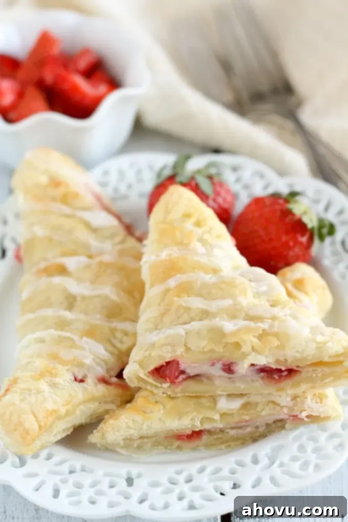 Two perfectly baked strawberry cream cheese turnovers artfully arranged on a white plate. One turnover is cut in half and stacked, proudly displaying its luscious strawberry and cream cheese interior. Fresh strawberries and forks are visible in the background.