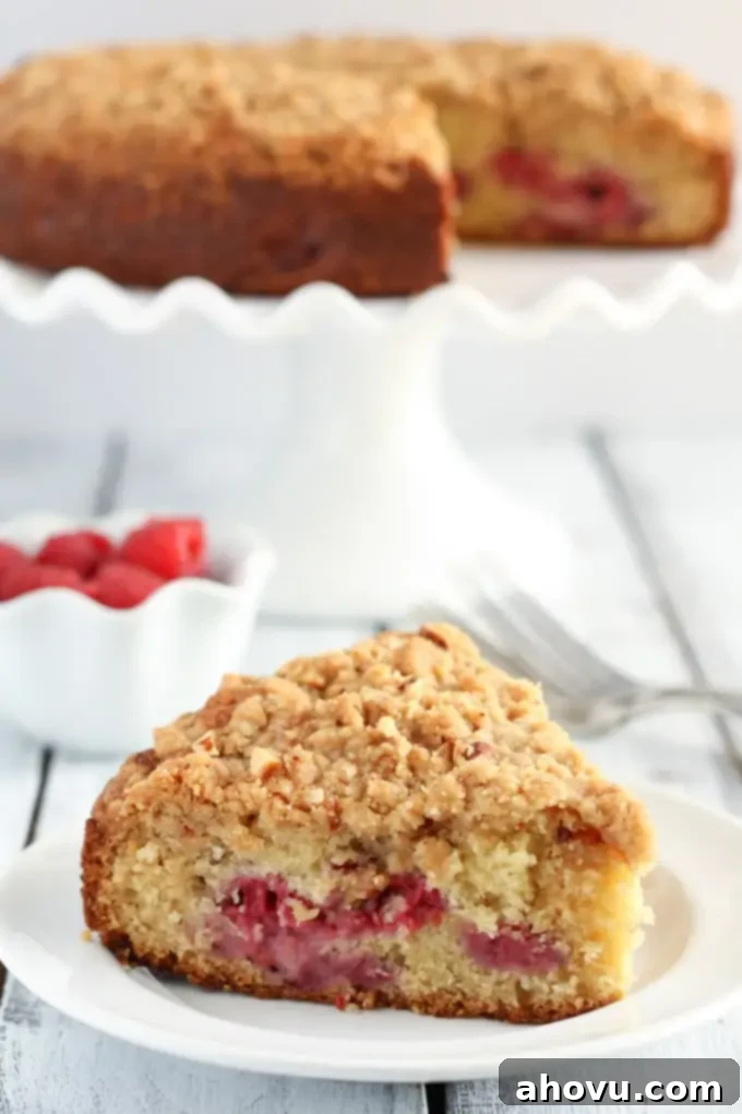 A slice of raspberry almond cake with crumb topping on a white plate. A dish of fresh raspberries and a cake on a cake stand rest in the background. 