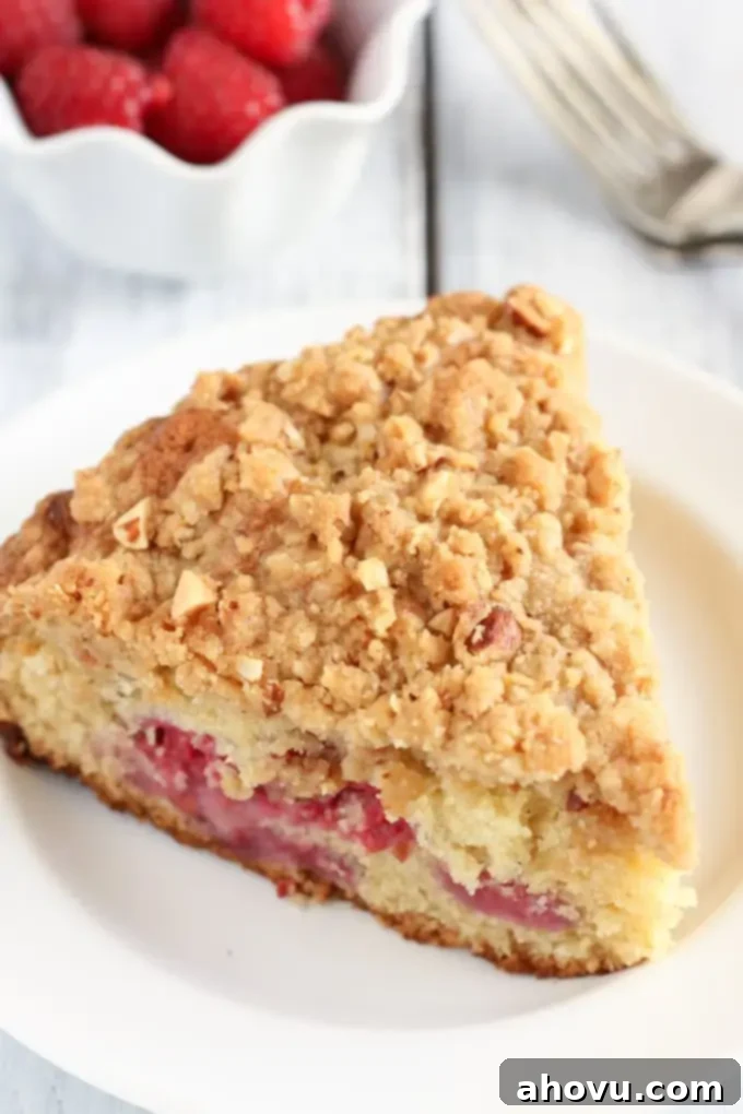 A slice of raspberry almond cake with crumb topping on a white plate. A bowl of fresh raspberries rest in the background. 