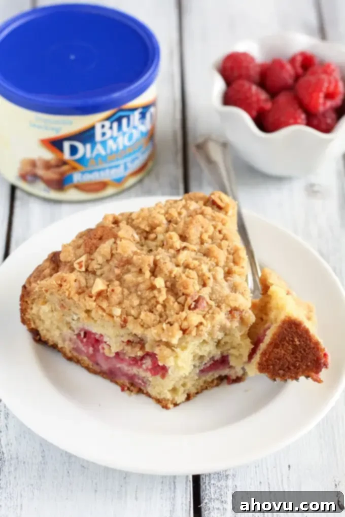 A slice of raspberry crumb cake on a white plate. A bowl of fresh raspberries and a container of almonds rest in the background. 