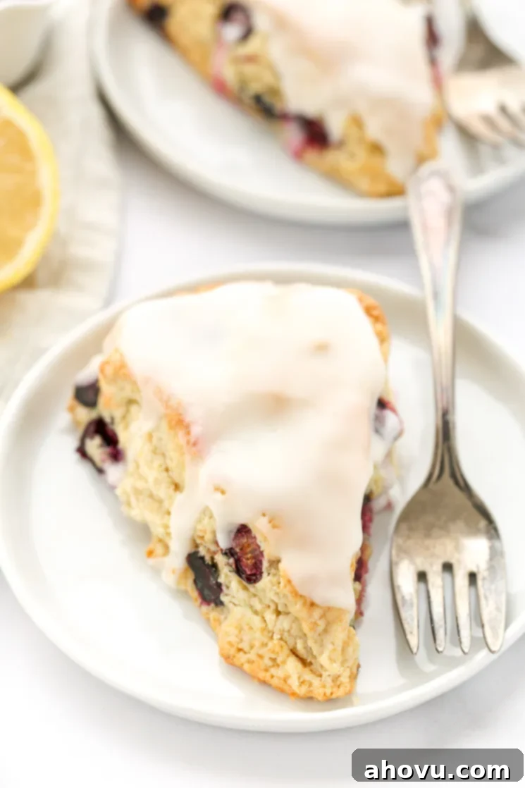A blueberry scone with lemon glaze on a white plate with another scone and sliced lemon in the background.