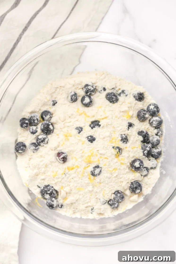 A glass bowl filled with the dry ingredients, blueberries, and lemon zest for scones and a light gray napkin on the side.