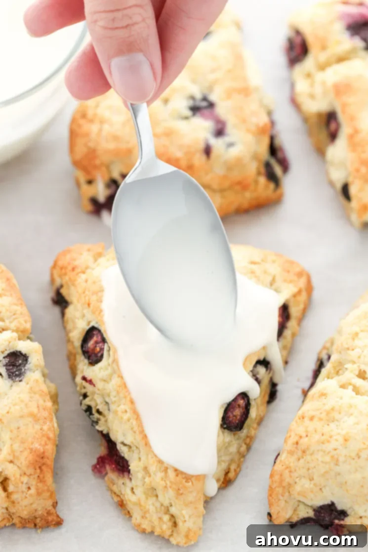 A closeup of the lemon blueberry scones on parchment paper being topped with the lemon glaze.