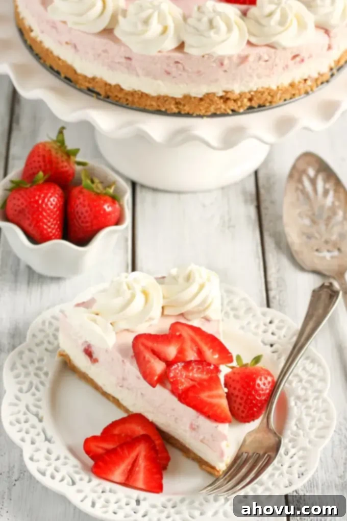 A slice of no bake strawberry cheesecake on a white dessert plate with a fork. A small dish of fresh strawberries and the remainder of the cheesecake rest in the background. 
