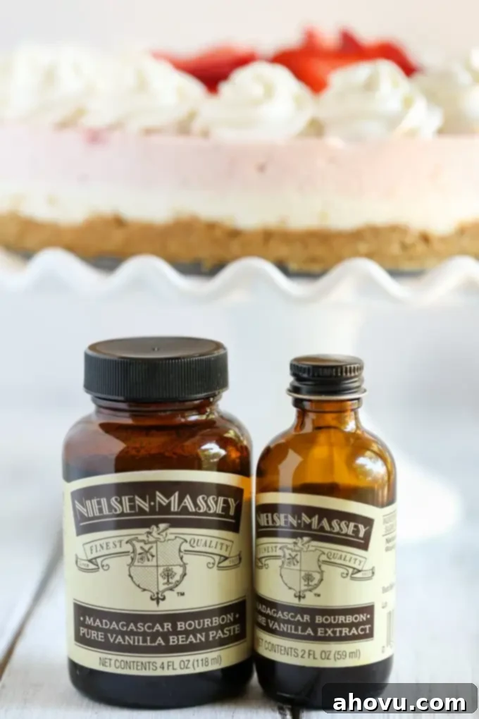 A bottle of vanilla bean paste and a bottle of vanilla extract. In the background is a fresh strawberry cheesecake on a cake stand. 