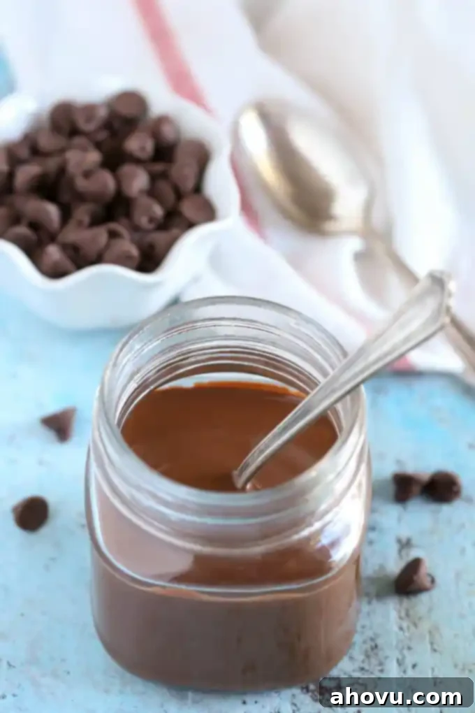 Homemade magic shell in a glass jar. A spoon is sticking out of the jar. A small dish of chocolate chips, a tea towel, and a spoon rest in the background. 