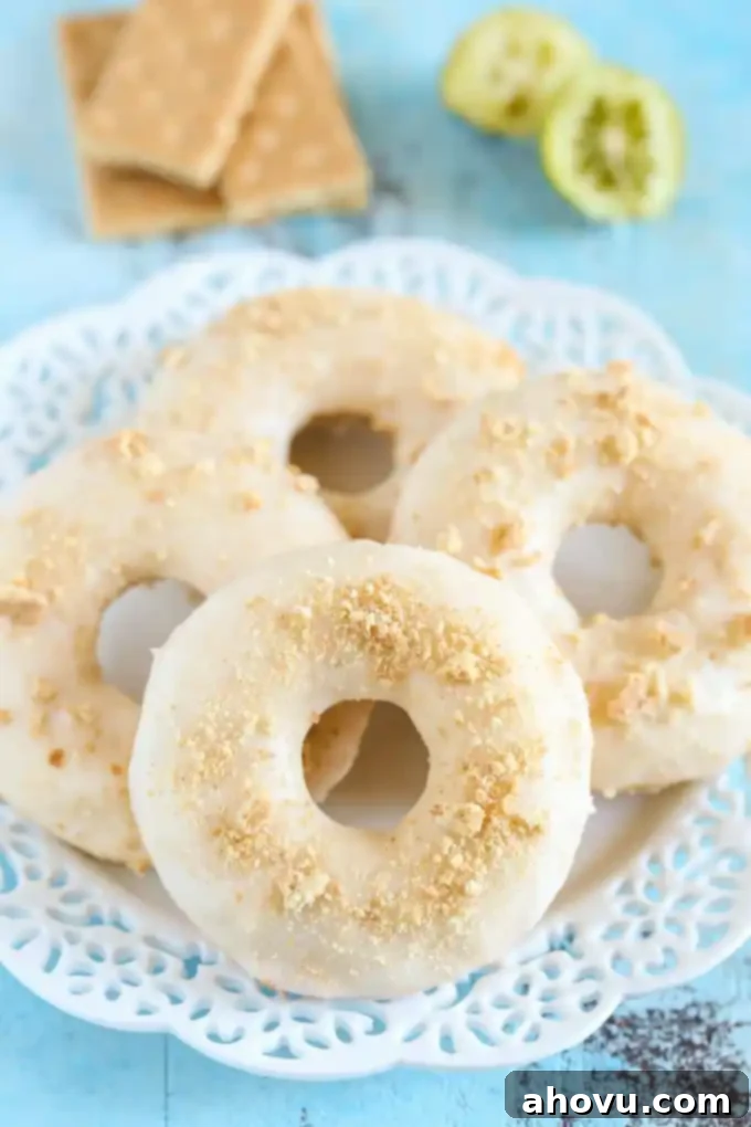 Four baked key lime donuts on a white plate. Graham crackers and lime halves rest in the background, creating a fresh and inviting scene.