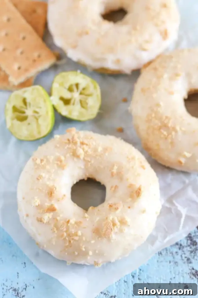 Three perfectly glazed key lime donuts arranged on parchment paper. Fresh lime halves and graham crackers are visible in the soft-focused background.