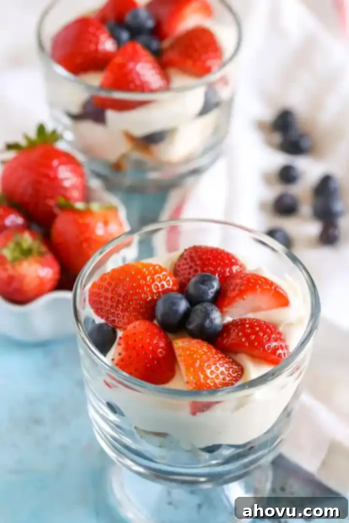 A small glass dish filled with angel food cake trifle, showing intricate layers. Another mini trifle and a dish of fresh strawberries are in the soft-focus background.