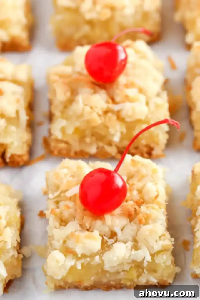 A top-down view of several rows of freshly sliced coconut pineapple bars, showcasing their golden-brown crumb topping and a vibrant, thick pineapple coconut filling. The two bars at the front are charmingly adorned with red maraschino cherries, adding a pop of color.