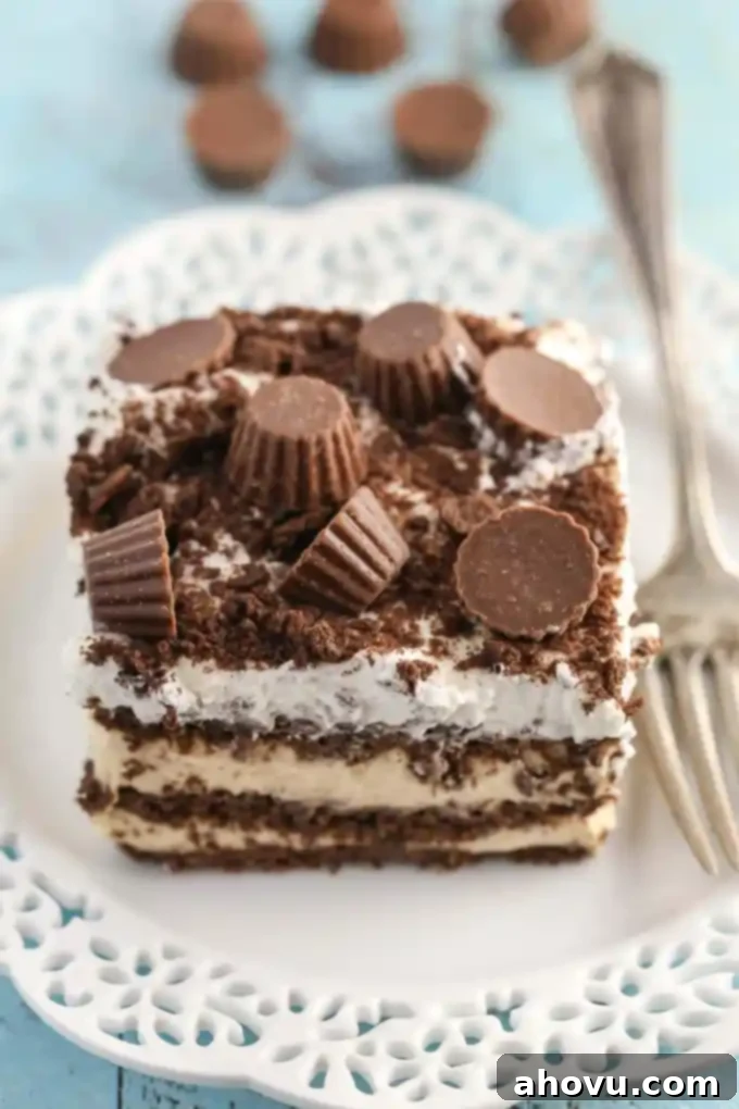 A perfectly sliced piece of peanut butter chocolate icebox cake, showing distinct layers of chocolate graham cracker and creamy peanut butter filling. It rests on a white plate with a fork, and scattered mini peanut butter cups are visible in the soft-focus background, inviting you to take a bite.