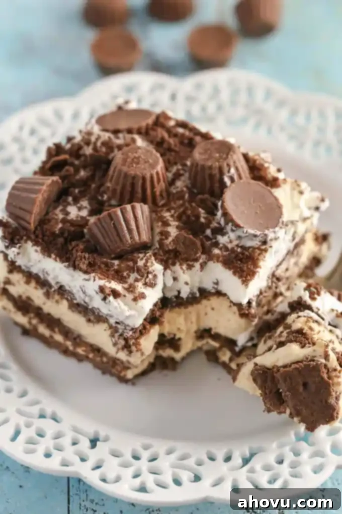 A close-up, angled shot of a slice of no-bake peanut butter chocolate icebox cake on a white plate. The layers are distinct, and the top is generously adorned with whipped cream and mini peanut butter cups, with a fork poised to scoop up a bite. The background is softly blurred.