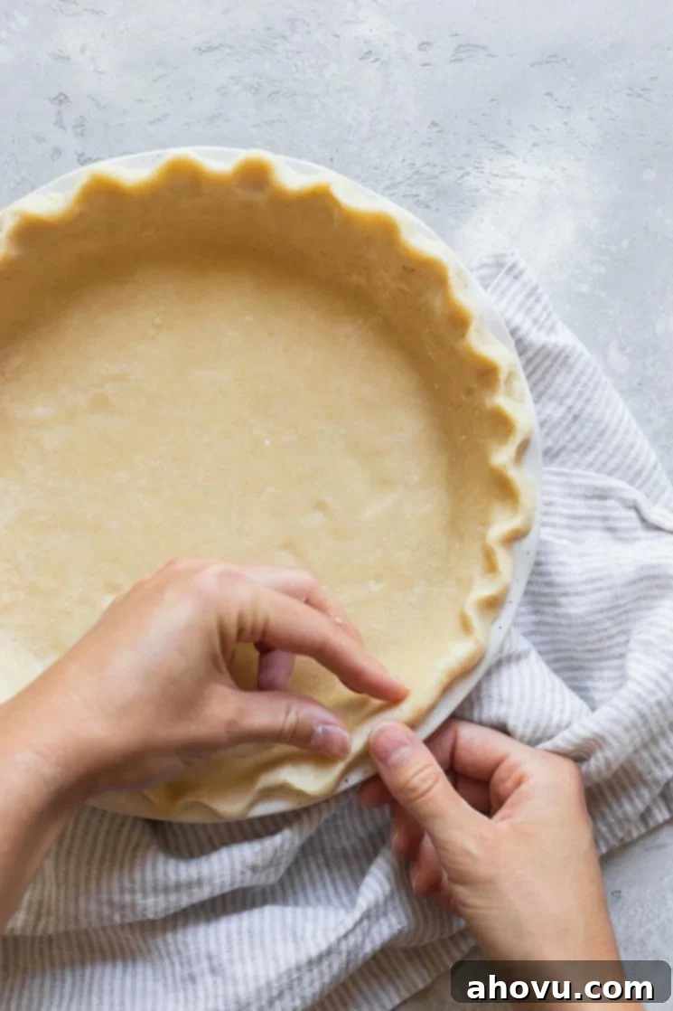 Close-up shot of hands carefully decorating the edges of an unbaked pie crust in a white pie dish, demonstrating a fluted crimp.
