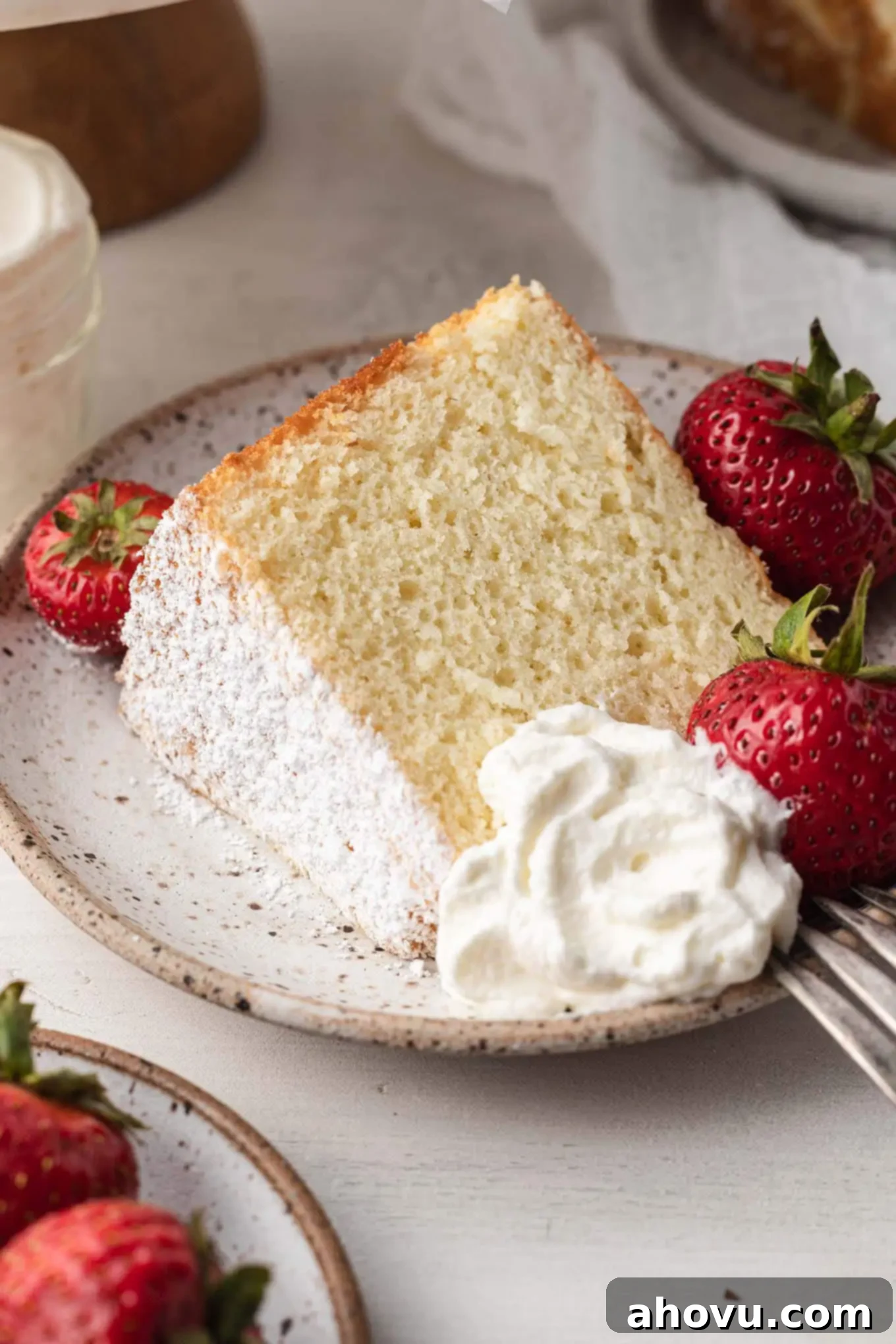A beautifully sliced chiffon cake on a white speckled plate, elegantly garnished with fresh strawberries and a dollop of homemade whipped cream, inviting indulgence.