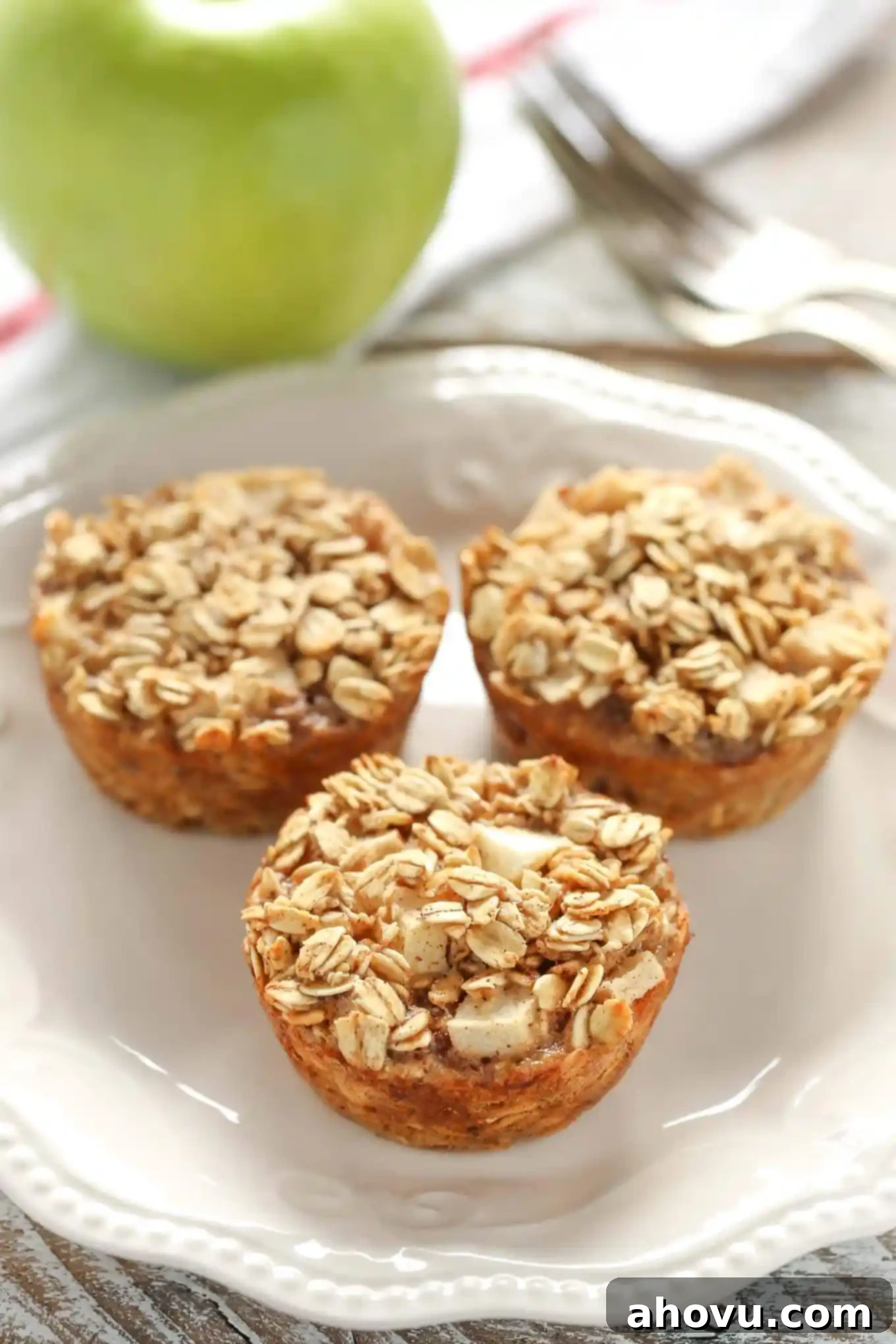 Three golden-brown baked apple oatmeal cups on a white plate. A fresh, crisp Granny Smith apple rests in the background on a textured tea towel, suggesting the key ingredient.