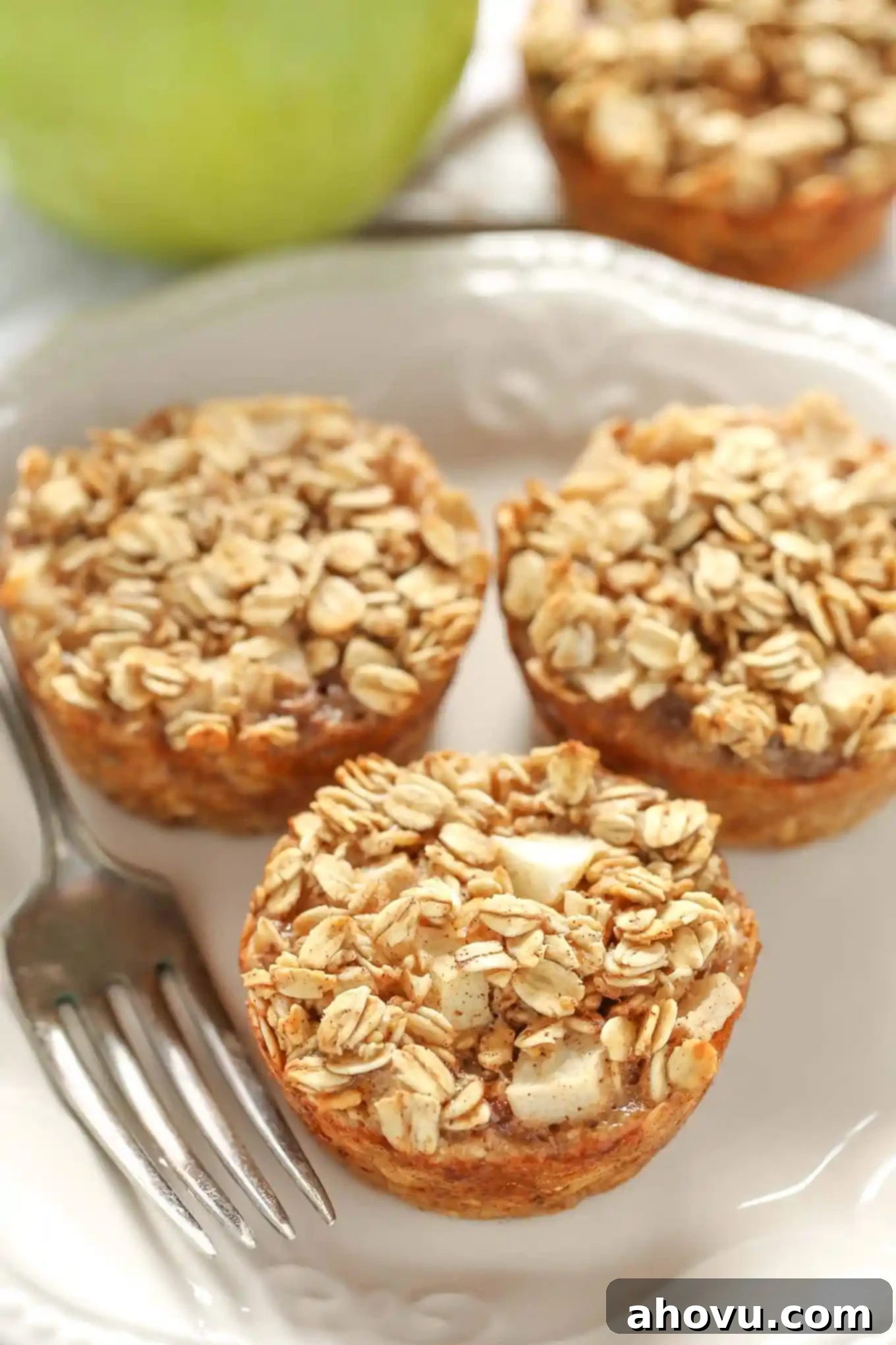 Three individual baked apple oatmeal cups arranged neatly on a white plate. A vibrant green Granny Smith apple adds a fresh touch in the soft-focused background, resting on a beige tea towel.