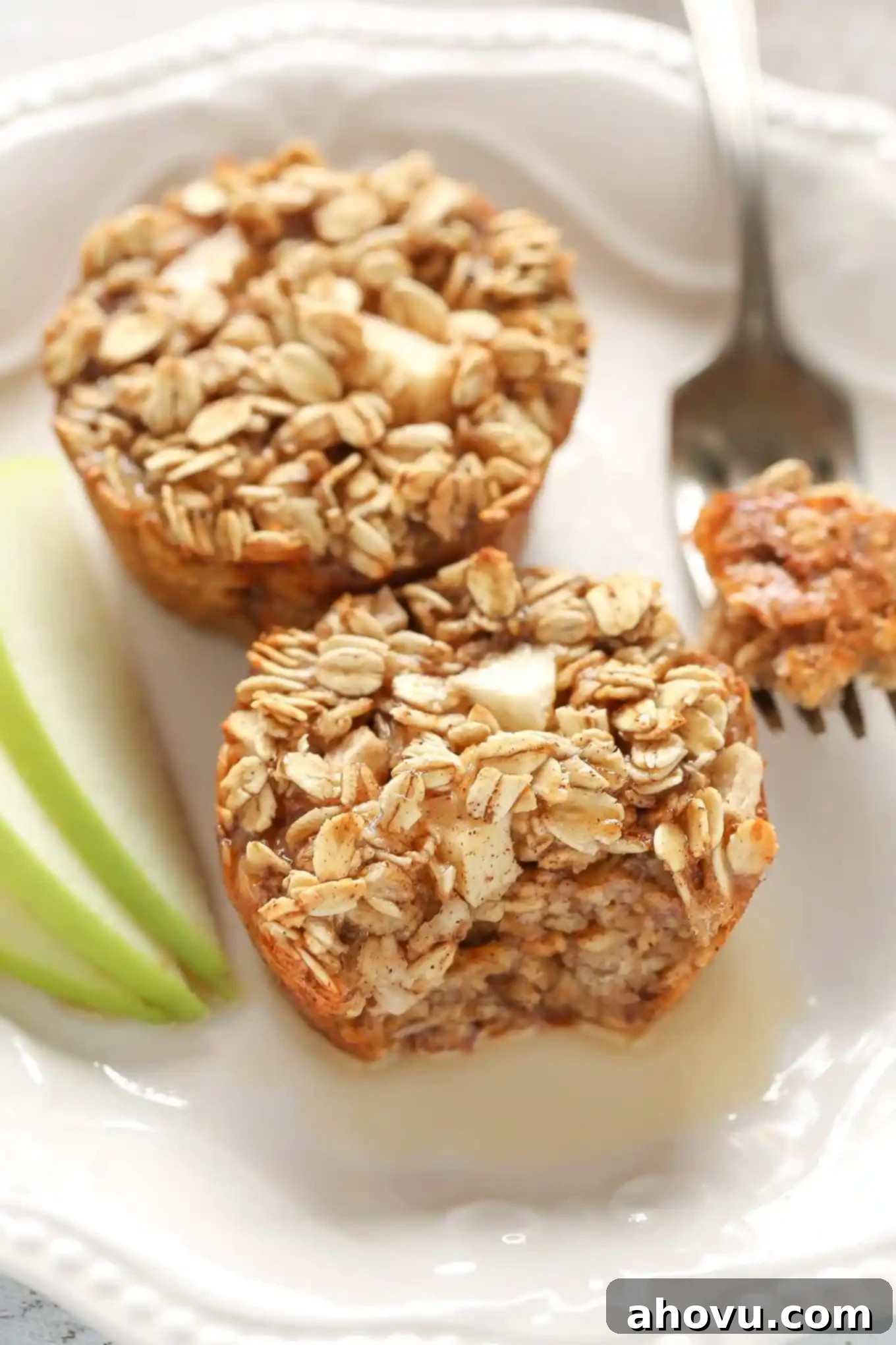 Two warm apple cinnamon baked oatmeal cups on a white plate, accompanied by a fork and decorative apple slices, highlighting the freshness of the ingredients.