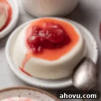 Silken Italian Indulgence 6 Panna cotta topped with homemade strawberry sauce on a speckled white plate. A spoon rests next to it.