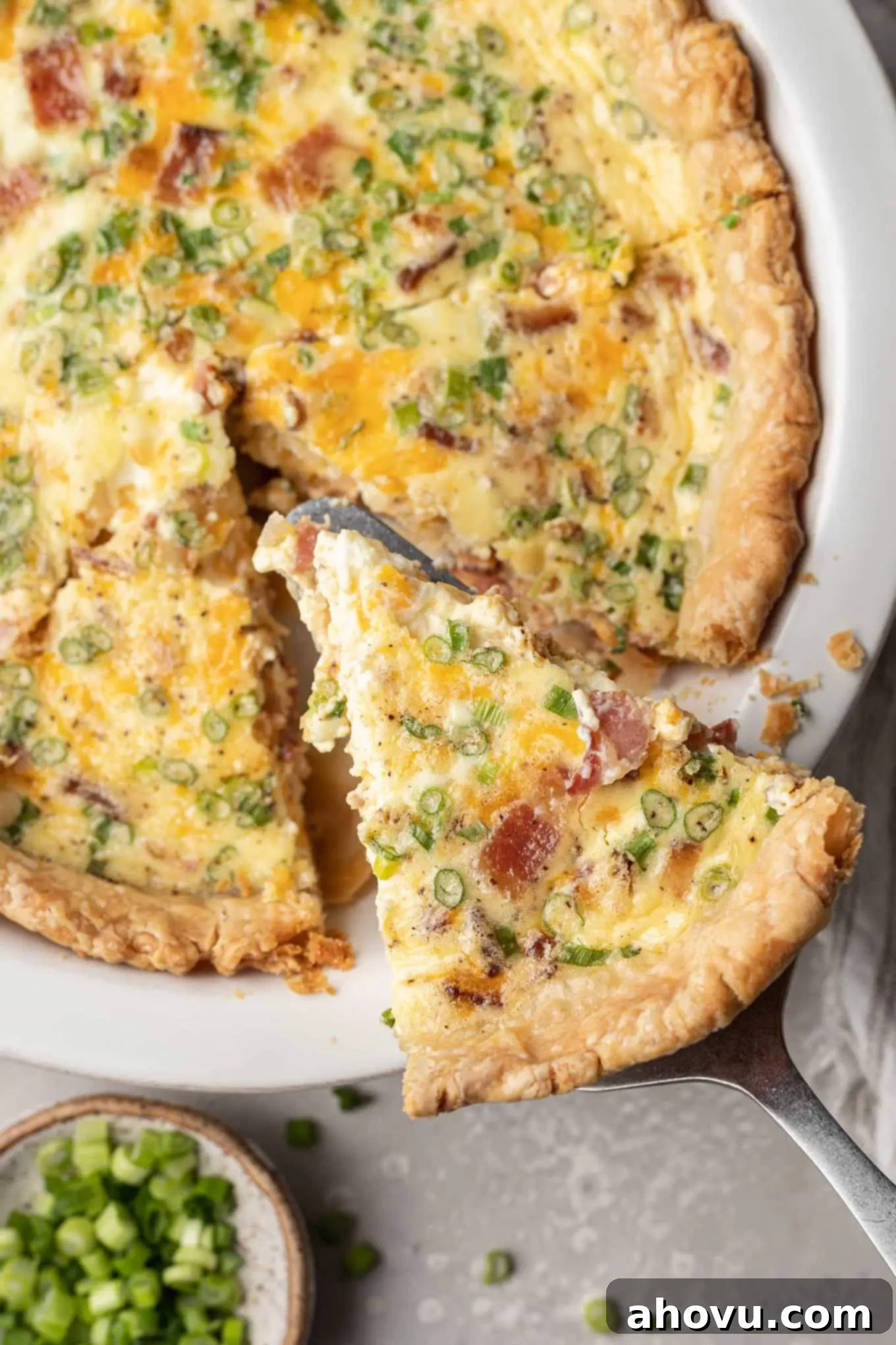 An overhead shot of a golden-brown slice of quiche being carefully removed from the white pie plate, showcasing its fluffy texture.