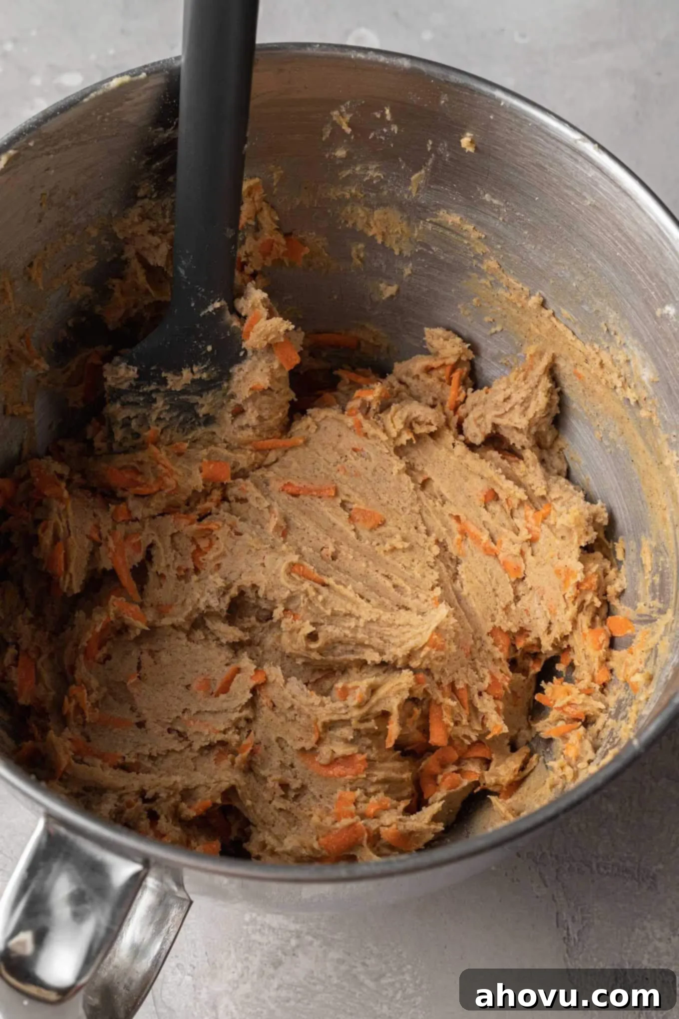 Raw carrot cake cookie dough in a metal mixing bowl with a rubber spatula, showing its texture before chilling and baking.