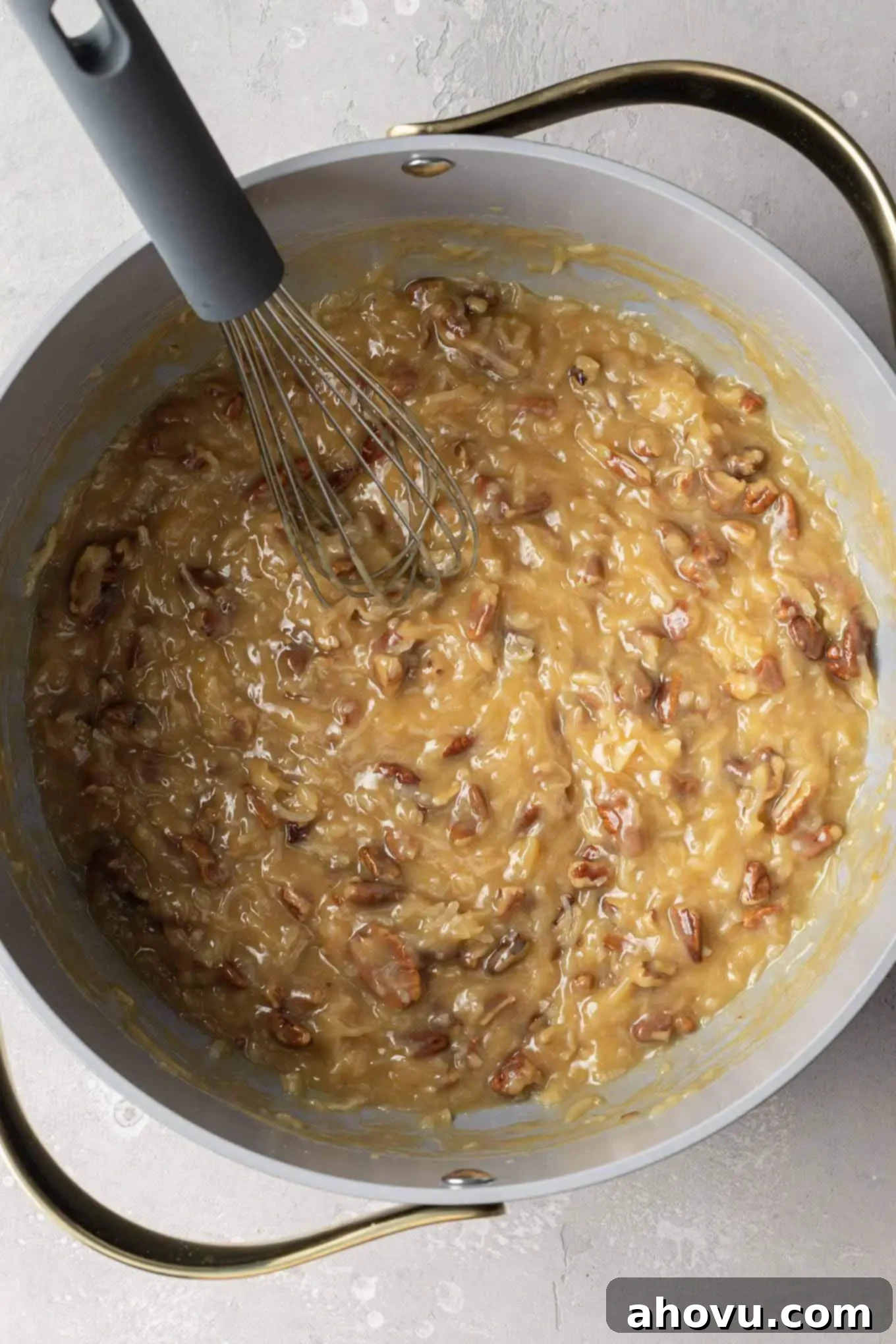 An overhead view of German chocolate cake frosting in a saucepan with a whisk, showing shredded coconut, pecans, and vanilla being mixed in.