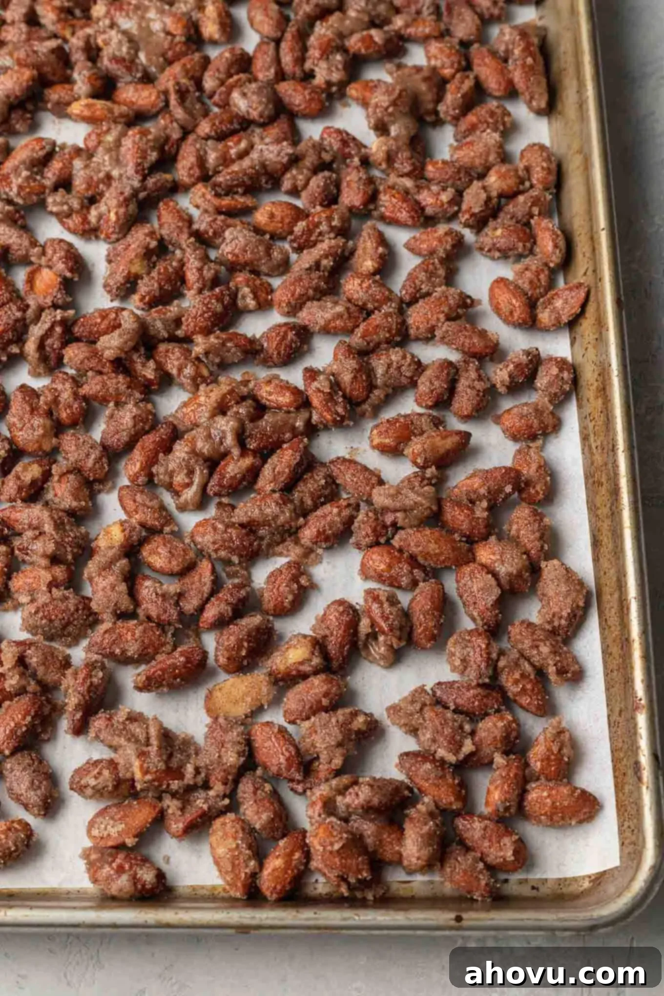 Unbaked candied almonds evenly spread in a single layer on a parchment paper-lined baking sheet, prepared for the oven.