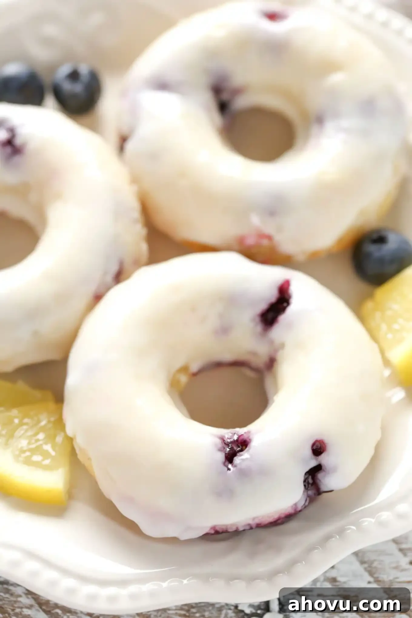 Zesty Lemon Blueberry Baked Donuts 2 Three lemon blueberry donuts topped with glaze on a white plate.