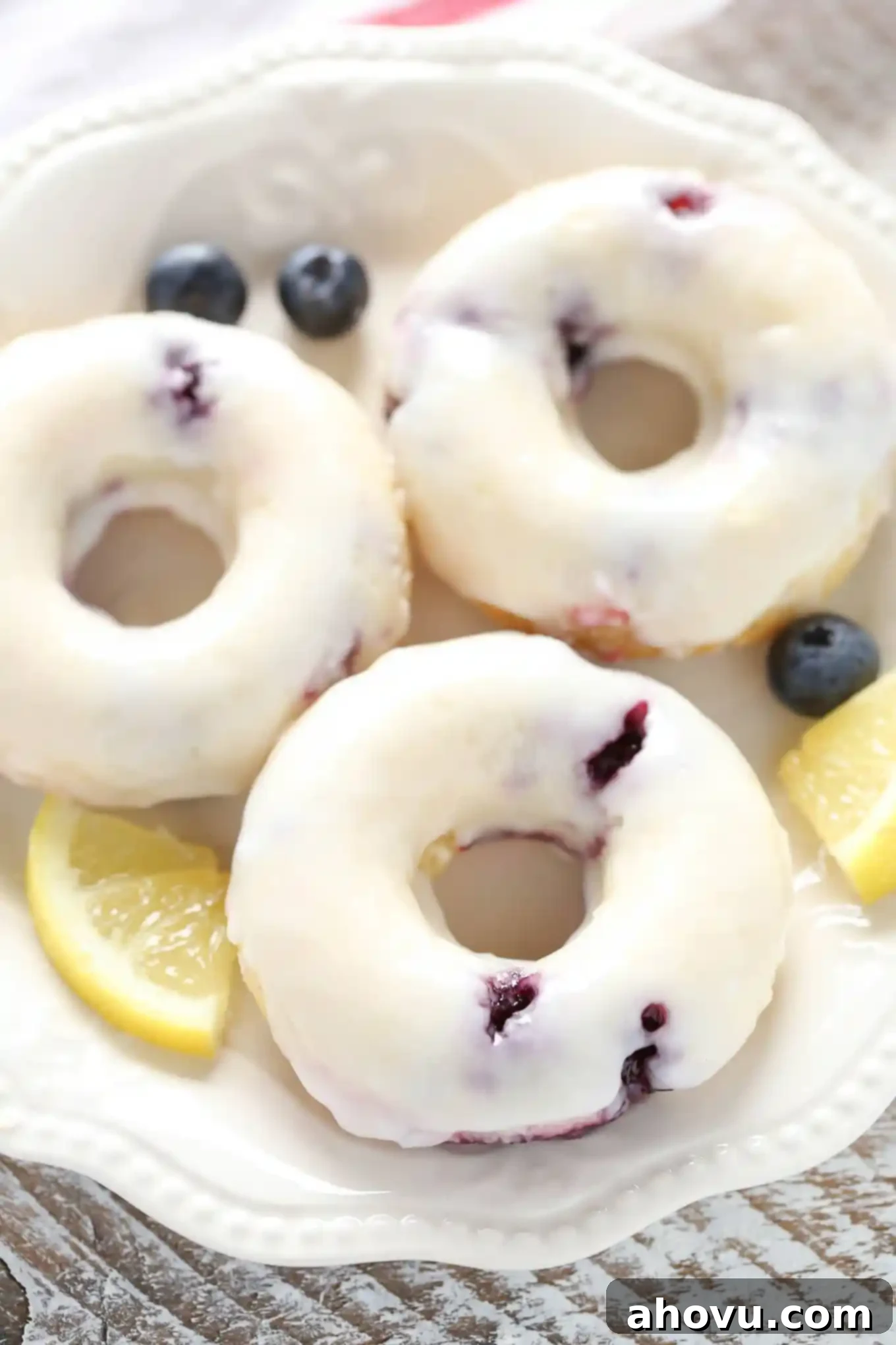 Zesty Lemon Blueberry Baked Donuts 4 Overhead view of three lemon blueberry donuts on a white plate. Small lemon wedges and fresh blueberries surround the plate.