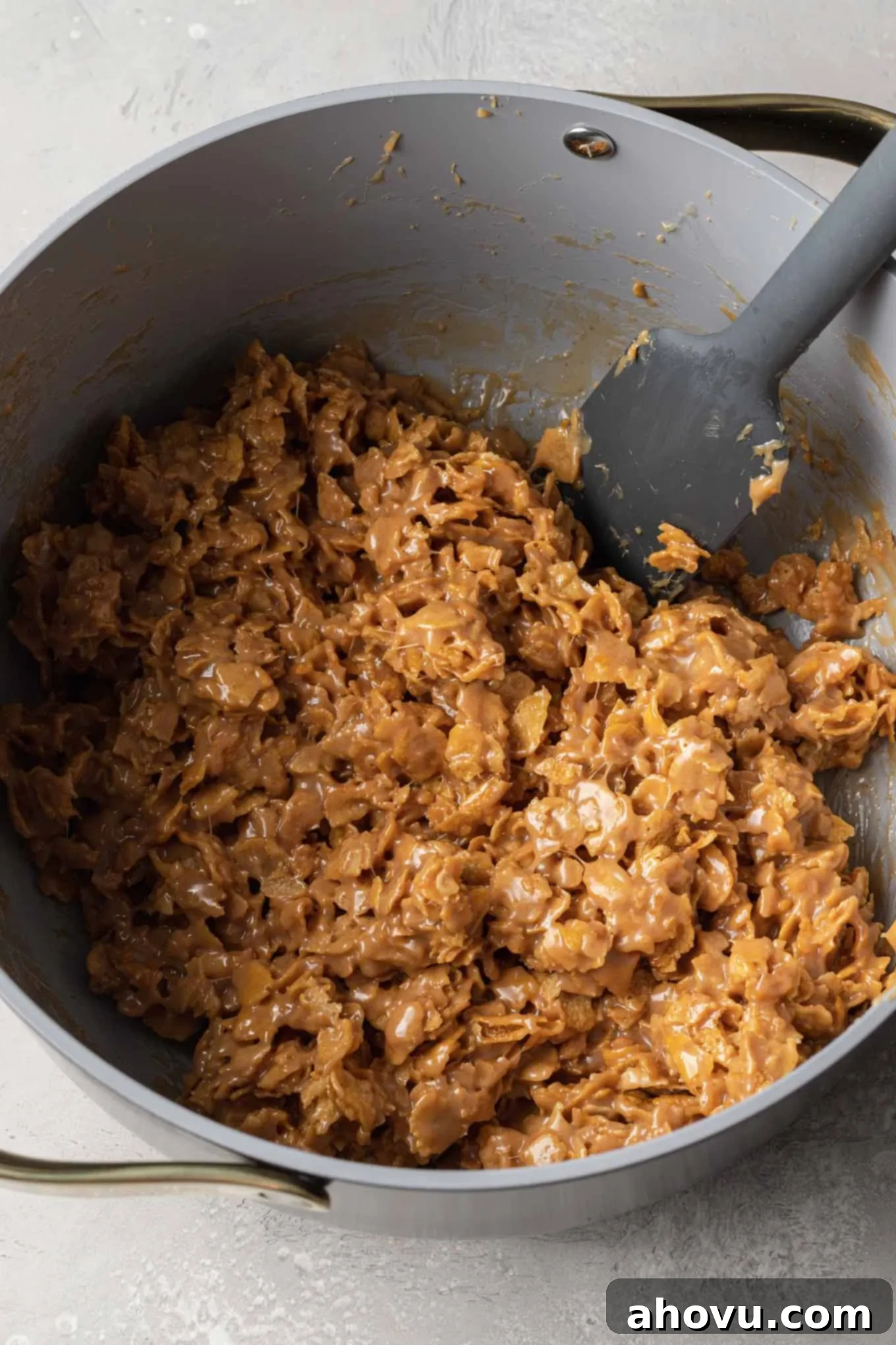 An overhead view of the crunchy cornflake cereal being gently folded into the peanut butter mixture in a saucepan, with a rubber spatula.