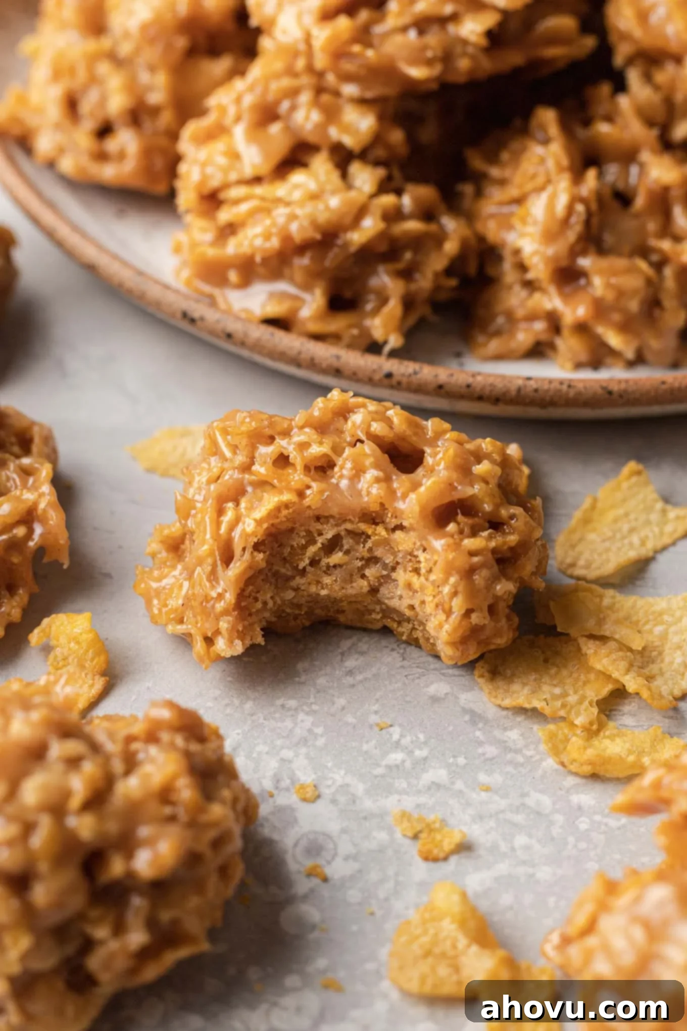A close-up view of a perfectly formed no-bake cornflake cookie with a bite missing, surrounded by more cookies on a serving plate.