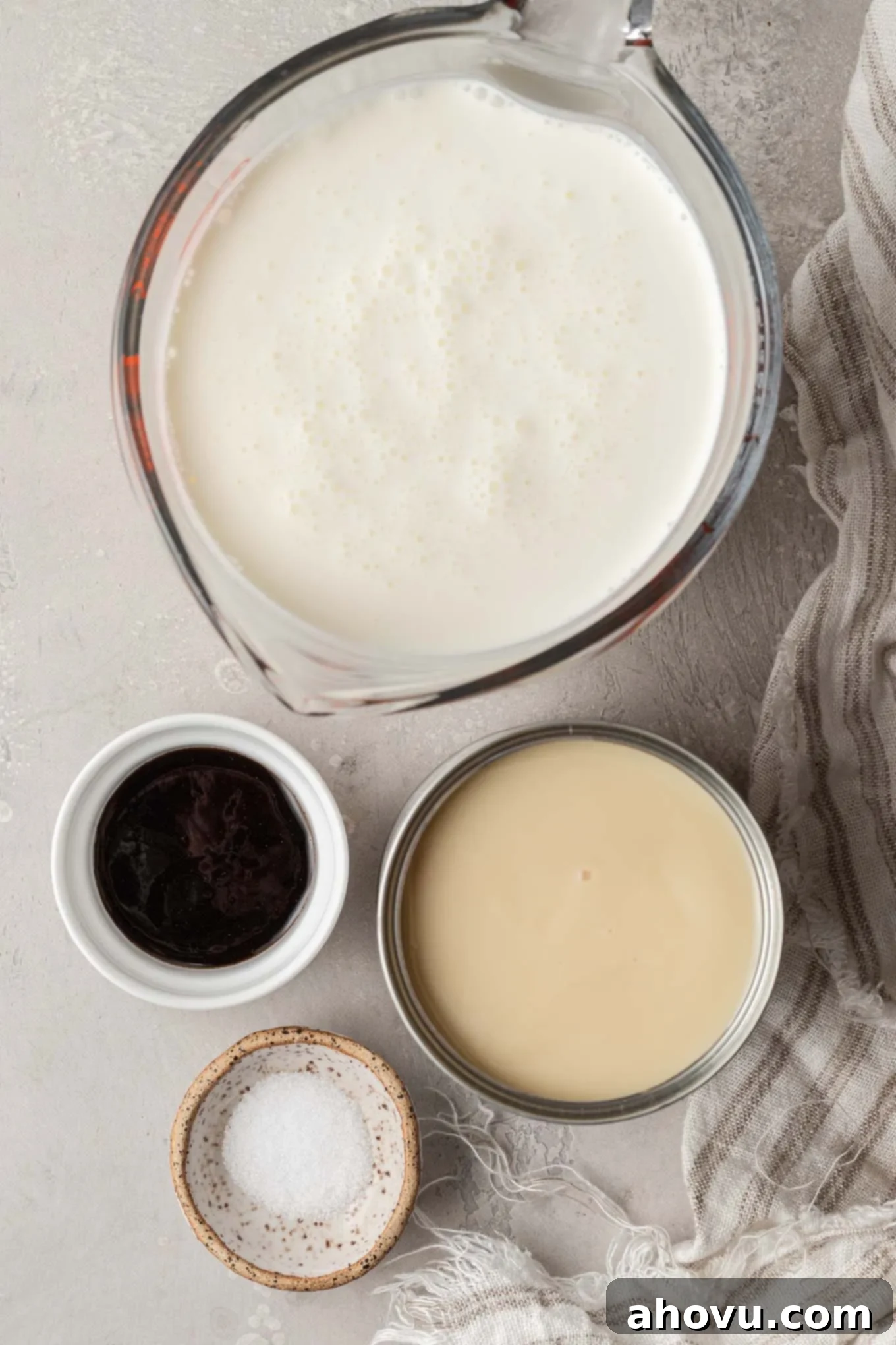 Overhead shot of essential no-churn ice cream ingredients: a can of sweetened condensed milk, a carton of heavy whipping cream, vanilla extract, and a pinch of salt on a white counter.