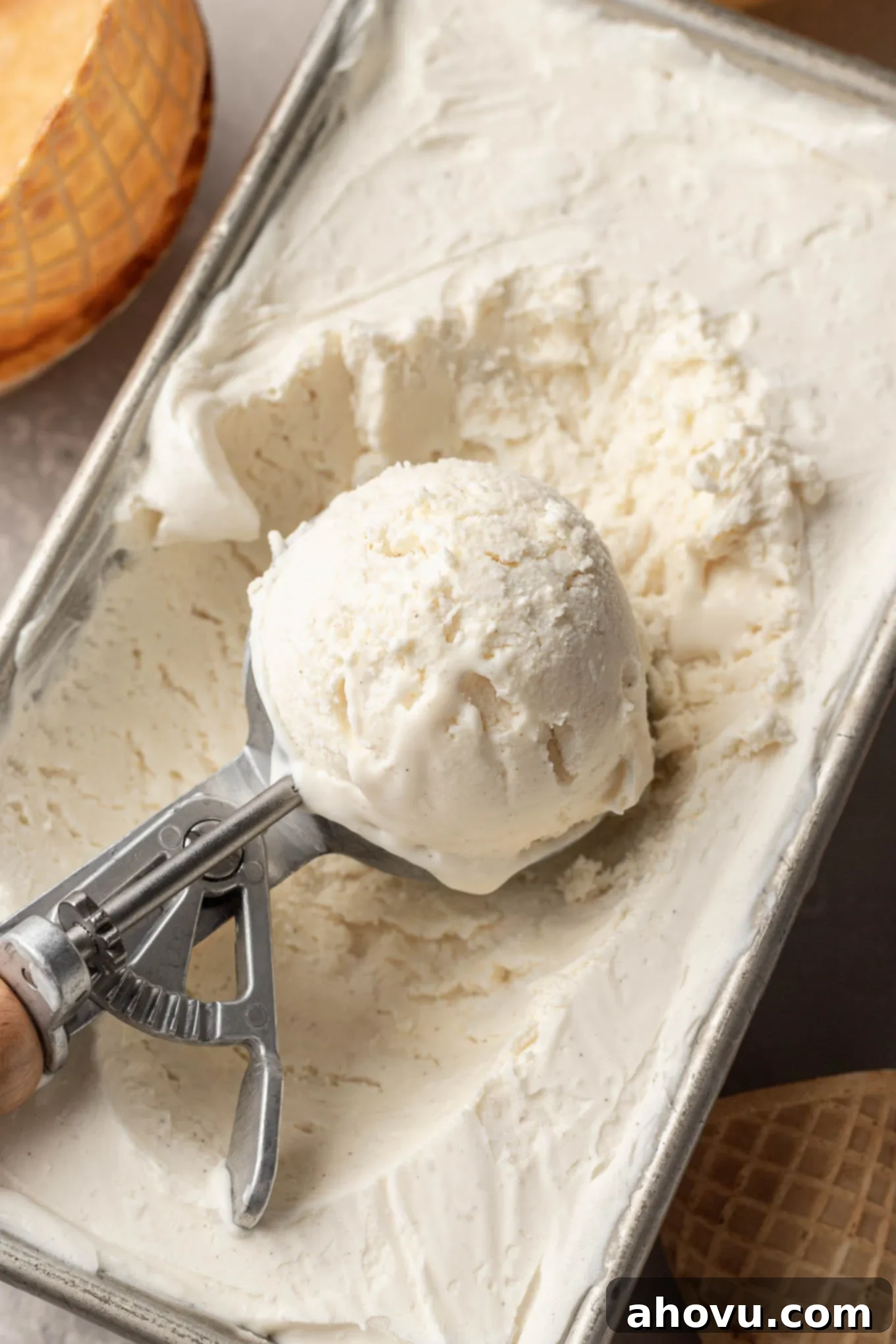 An ice cream scoop poised above a pan of freshly frozen homemade no-churn vanilla ice cream, ready to serve.