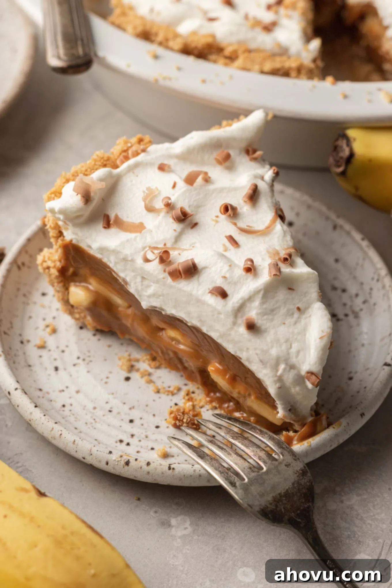 A beautiful slice of homemade banoffee pie on a speckled white dessert plate, showcasing its distinct layers of crust, dulce de leche, bananas, and whipped cream. 